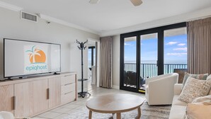 A modern living room with a television displaying 'epihost', a ceiling fan, a balcony with a view of the ocean, and a dining area with a round table. - VillaSolare-Sandestin: 9th-Floor Gulf-Front+Views (Miramar Beach)