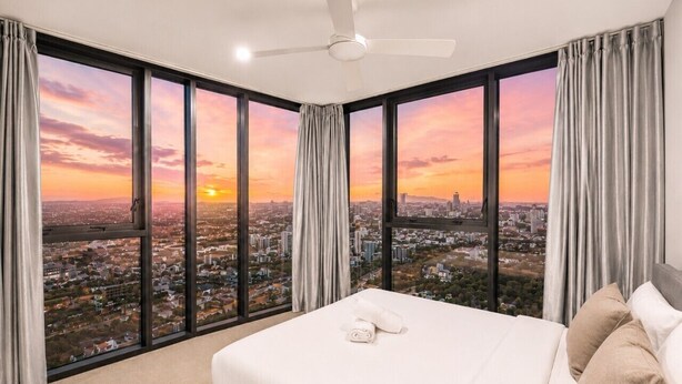 A hotel room with a large bed, a ceiling fan, and a city view. - City Lights Views – 2BR Stay (South Brisbane)