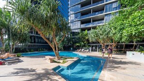 A swimming pool with lounge chairs, palm trees, and a modern building in the background. - City Lights Views – 2BR Stay (South Brisbane)