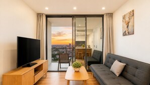 A modern living room with a flat-screen TV, a grey sofa, a wooden coffee table, and a city view through a glass door. - City Lights Views – 2BR Stay (South Brisbane)