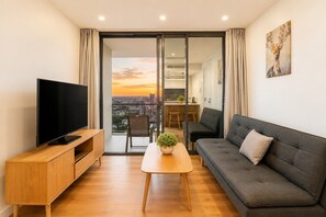 A modern living room with a flat-screen TV, a grey sofa, a wooden coffee table, and a city view through a glass door.