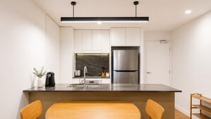 A modern kitchen with white cabinets, a stainless steel refrigerator, a marble backsplash, and a wooden dining table with chairs. - City Lights Views – 2BR Stay (South Brisbane)