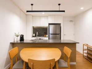 A modern kitchen with white cabinets, a stainless steel refrigerator, a marble backsplash, and a wooden dining table with chairs.
