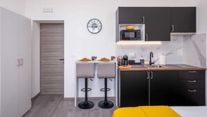 Cocina compacta con microondas, lavaplatos y armarios negros. Hay dos taburetes de bar y una mesita con platos y vasos.