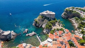 Vista aérea de un pueblo costero con un edificio histórico destacado sobre un promontorio rocoso, aguas azules cristalinas y un puerto deportivo con barcos.