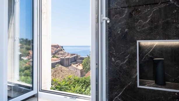 Una habitación con un ventanal grande que ofrece vistas a un paisaje urbano costero, una pared de mármol y un estante con un objeto cilíndrico.