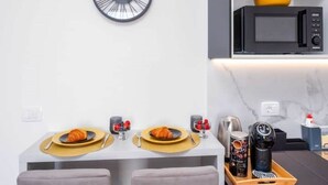 Una cocina moderna con microondas, un reloj y un comedor con dos sillas y una mesa dispuesta con platos, vasos y un croissant.