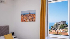 Habitación de hotel con cama, mesita de noche, un cuadro de una ciudad costera y ventana con vistas a un acantilado y al mar.