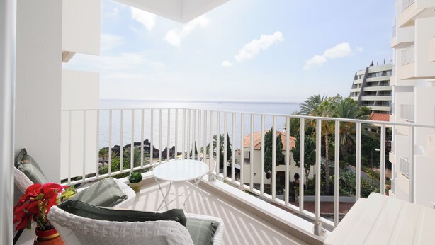 A balcony with a white railing, a white table, and a white sofa. There is a potted plant with red flowers on the left side. - Apartamentos do Mar - Pikis Flat (Funchal)