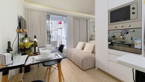 A modern kitchen with a built-in oven and microwave, a dining area with a table set for two, and a living area with a sofa. - Apartamentos do Mar - Pikis Flat (Funchal)