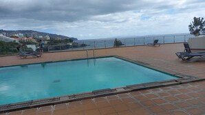 A swimming pool with tiled deck and lounge chairs, overlooking a coastal area with buildings and hills. - Apartamentos do Mar - Pikis Flat (Funchal)