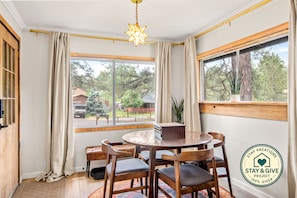 A room with a round wooden table, four chairs, a potted plant, and a window with a view of trees and a vehicle outside.