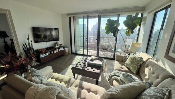 A spacious living room with a large sofa, a coffee table, and a TV on a stand. There are several plants and a balcony with a view of the city. - Urban Oasis with Sweeping Views - Walk to Gaslamp, Little Italy, Balboa Park (San Diego)