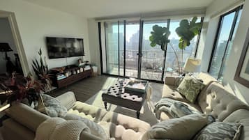 Un salon spacieux avec un grand canapé, une table basse et un téléviseur posé sur un meuble. On y trouve également plusieurs plantes et un balcon offrant une vue sur la ville.