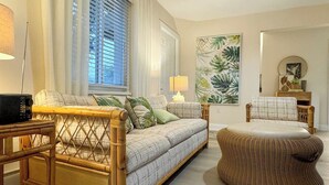Una sala de estar con un sofá, un sillón de mimbre, una lámpara y un cuadro grande con una hoja tropical en la pared.