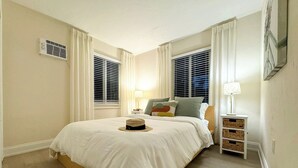 Un dormitorio con una cama grande, dos mesitas de noche con lámparas, una ventana con persianas y un respiradero en la pared.