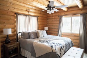 Un dormitorio de cabaña de madera con una cama, lámparas de noche, ventilador de techo y ventanas amplias.