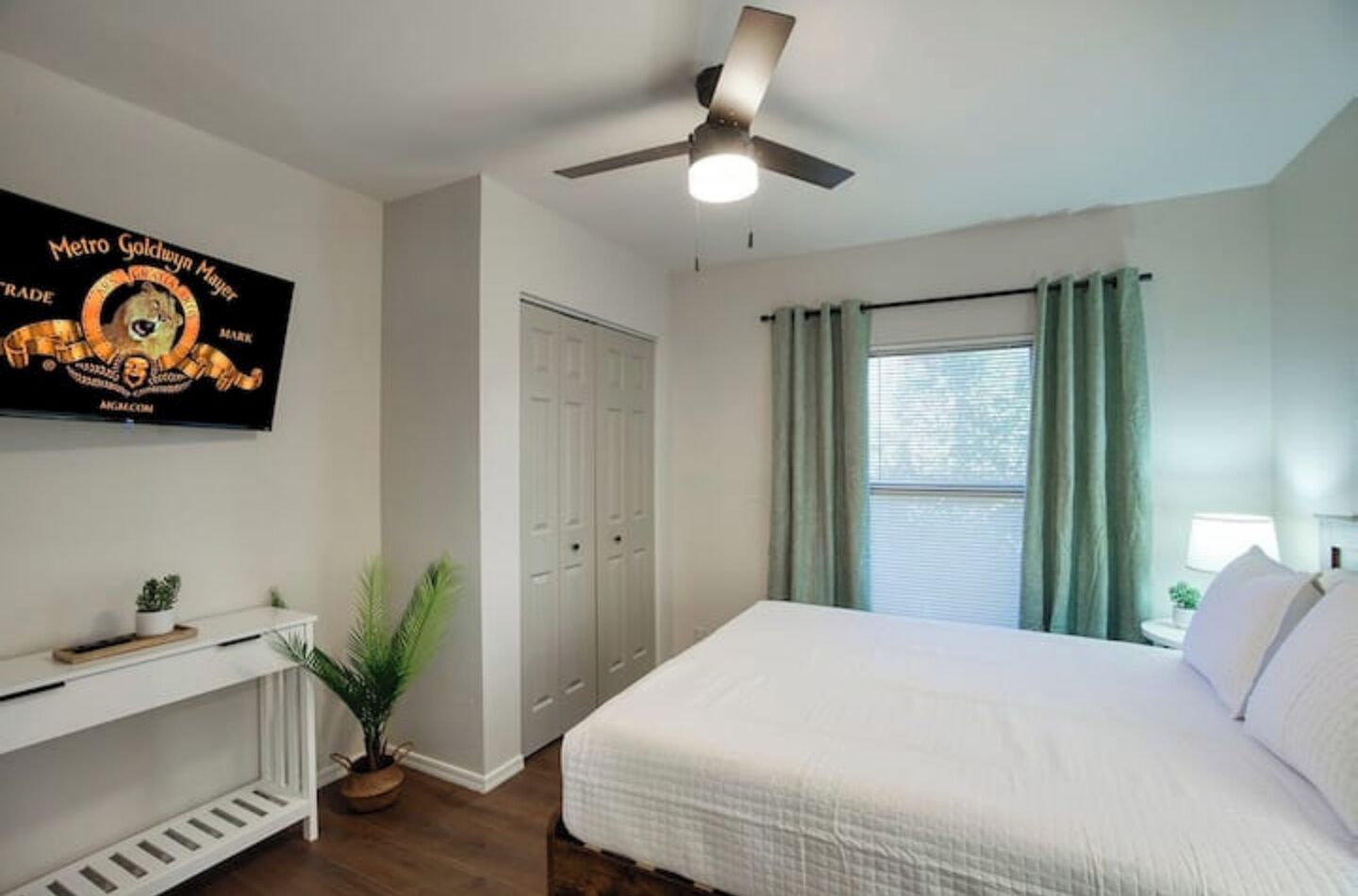 Un dormitorio con cama, ventilador de techo, televisor montado en la pared, armario y ventana con cortinas.