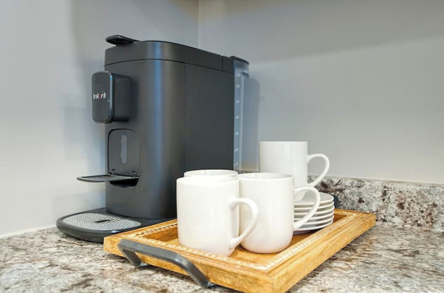 A coffee maker on a granite countertop with two white mugs and a stack of white plates on a wooden tray.