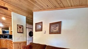 Una sala de estar acogedora con un sofá marrón, cojines estampados, techo de madera y una cocina al fondo.