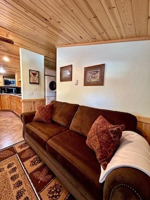 A cozy living room with a brown sofa, patterned cushions, a wooden ceiling, and a kitchen area in the background.