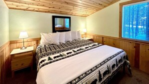 Un dormitorio con techo de madera, una cama con colcha estampada, dos mesitas de noche con lámparas, una ventana con persianas y un espejo sobre la cama.