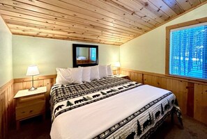 A bedroom with a wooden ceiling, a bed with a patterned bedspread, two bedside tables with lamps, and a window with a view of trees.