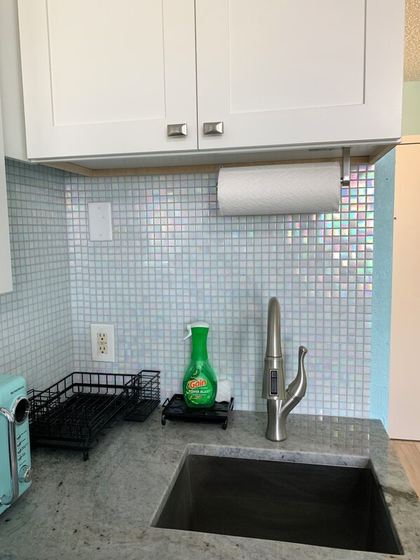 Una cocina con un lavamanos de acero inoxidable, un pulverizador verde, un dispensador de toallas de papel y una cesta de alambre negra.