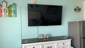 A room with a flat-screen TV mounted on the wall, a white dresser with drawers, and a refrigerator.