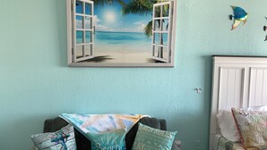 A room with a beach-themed wall art, a grey sofa with beach-themed cushions, and a white bed with a patterned bedspread.