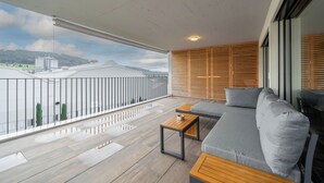 Un balcon doté d’un plancher en bois, d’un canapé gris agrémenté de coussins, d’une petite table en bois et offrant une vue sur des bâtiments et des collines.