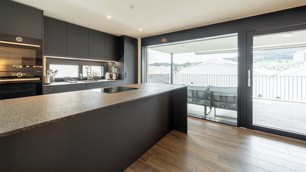 Une cuisine moderne avec des meubles de couleur sombre, un plan de travail en granit et une grande fenêtre donnant sur un balcon.
