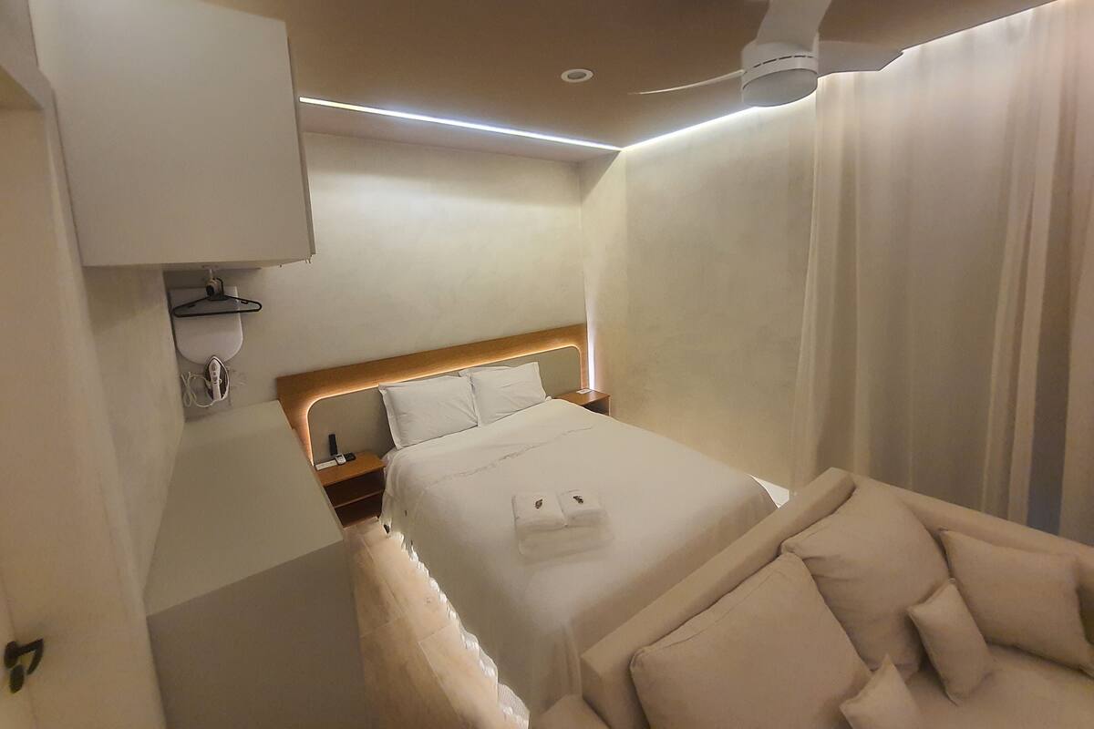 Une chambre d’hôtel avec un lit, une table de chevet, un miroir, un ventilateur de plafond et une fenêtre avec des rideaux.