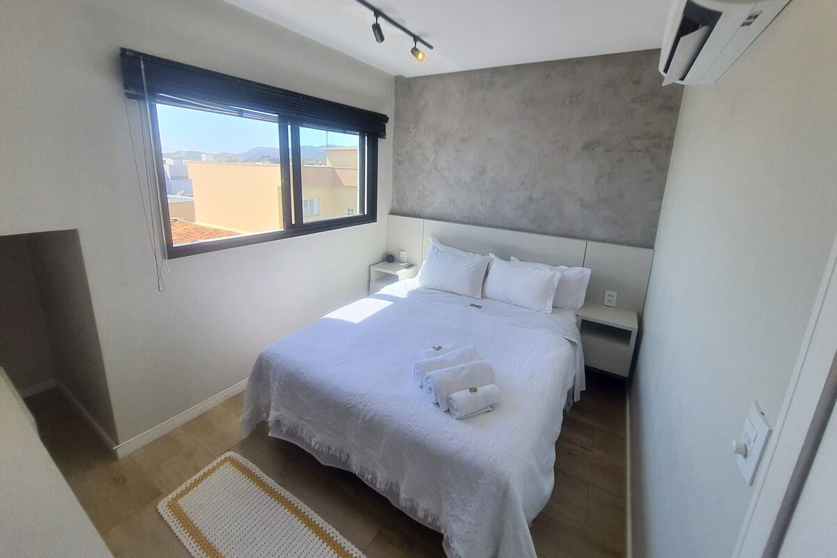 Une chambre avec un grand lit, une fenêtre, une table de chevet et un climatiseur fixé au mur.