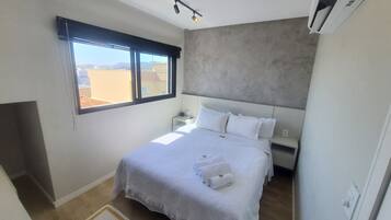 A bedroom with a large bed, a window, a bedside table, and a wall-mounted air conditioner.