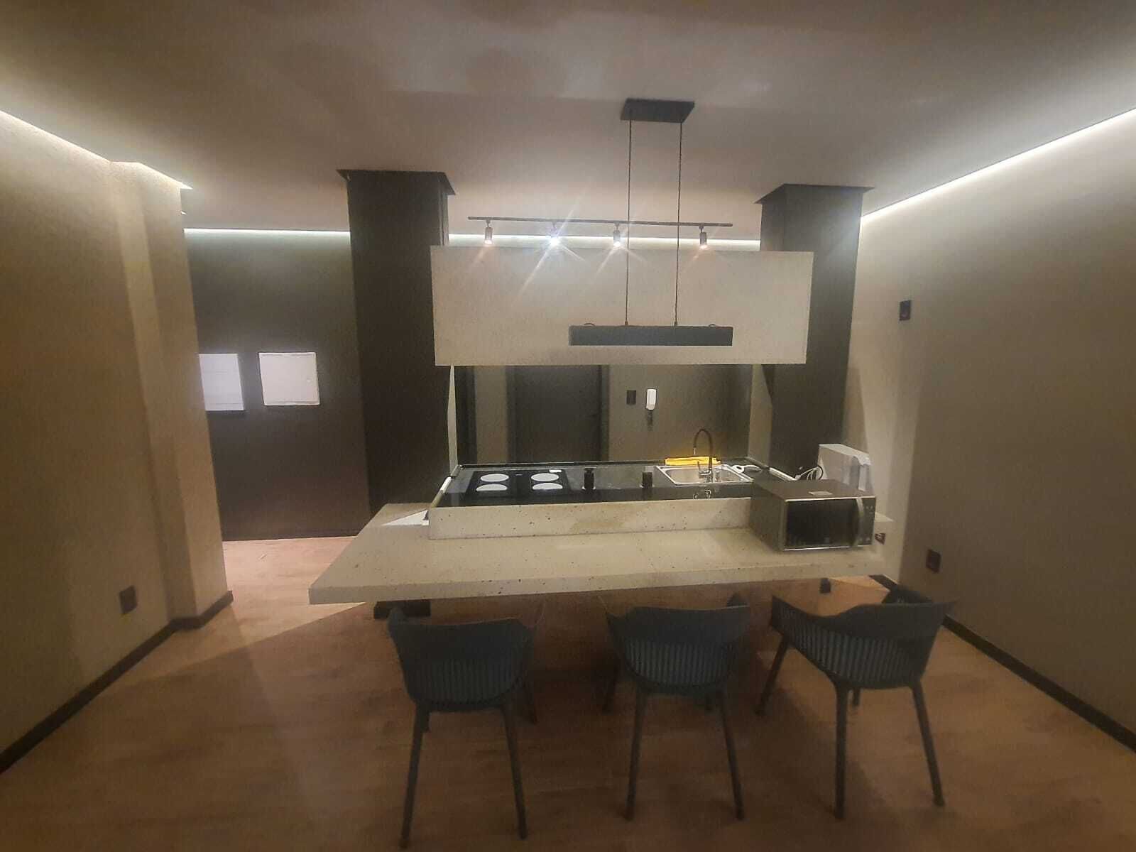 Uma cozinha moderna com uma ilha central, forno embutido e uma pia. Há três cadeiras ao redor da ilha.