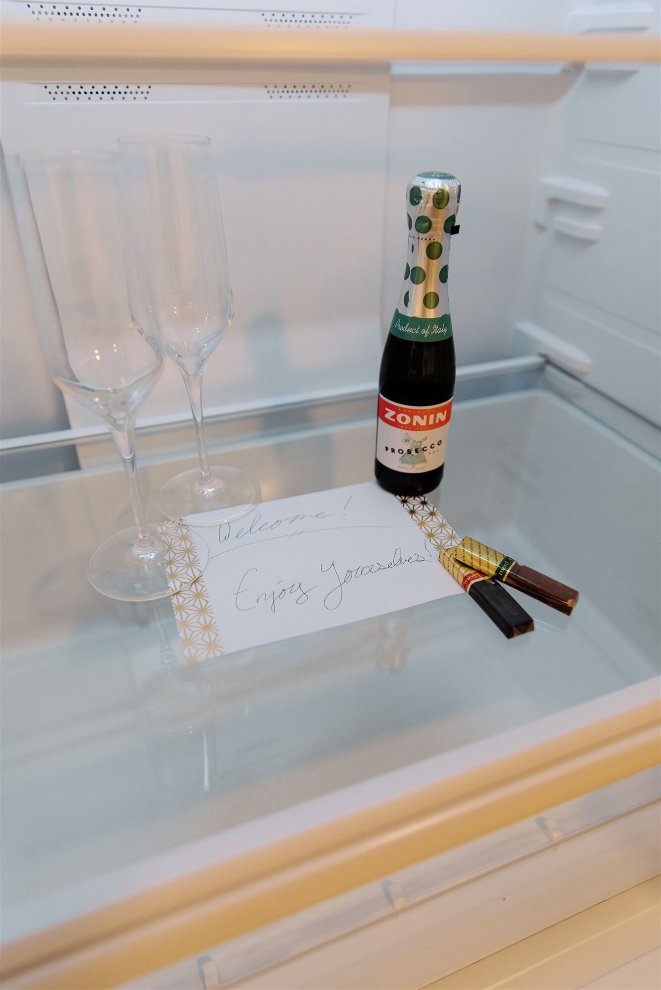 Un refrigerador con una botella de Zonin Prosecco, dos copas de vino vacías y una nota que dice '¡Bienvenidos! ¡Disfruten!'