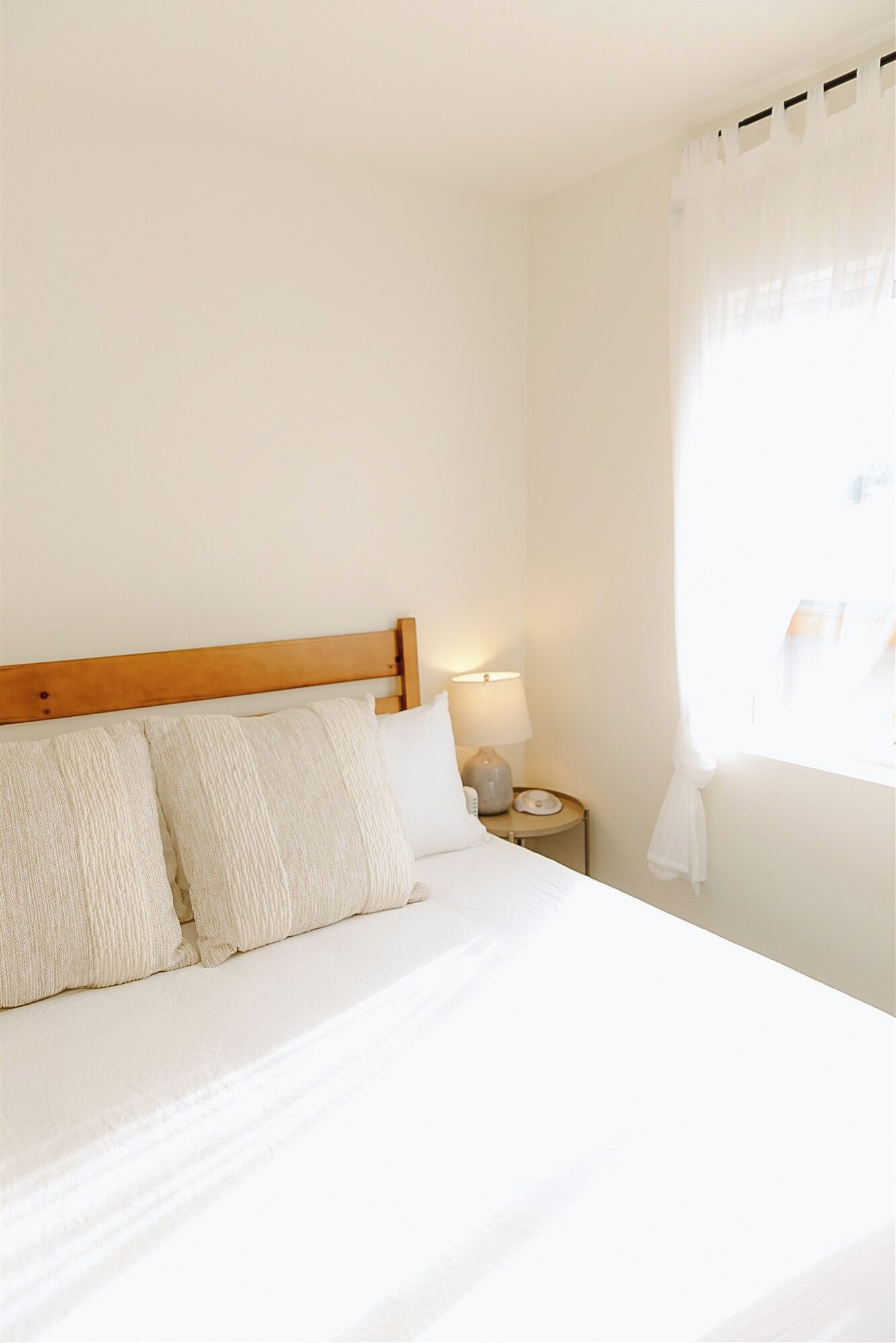 Una cama bien hecha con cabecera de madera, una lámpara de noche y cortinas translúcidas en una ventana.