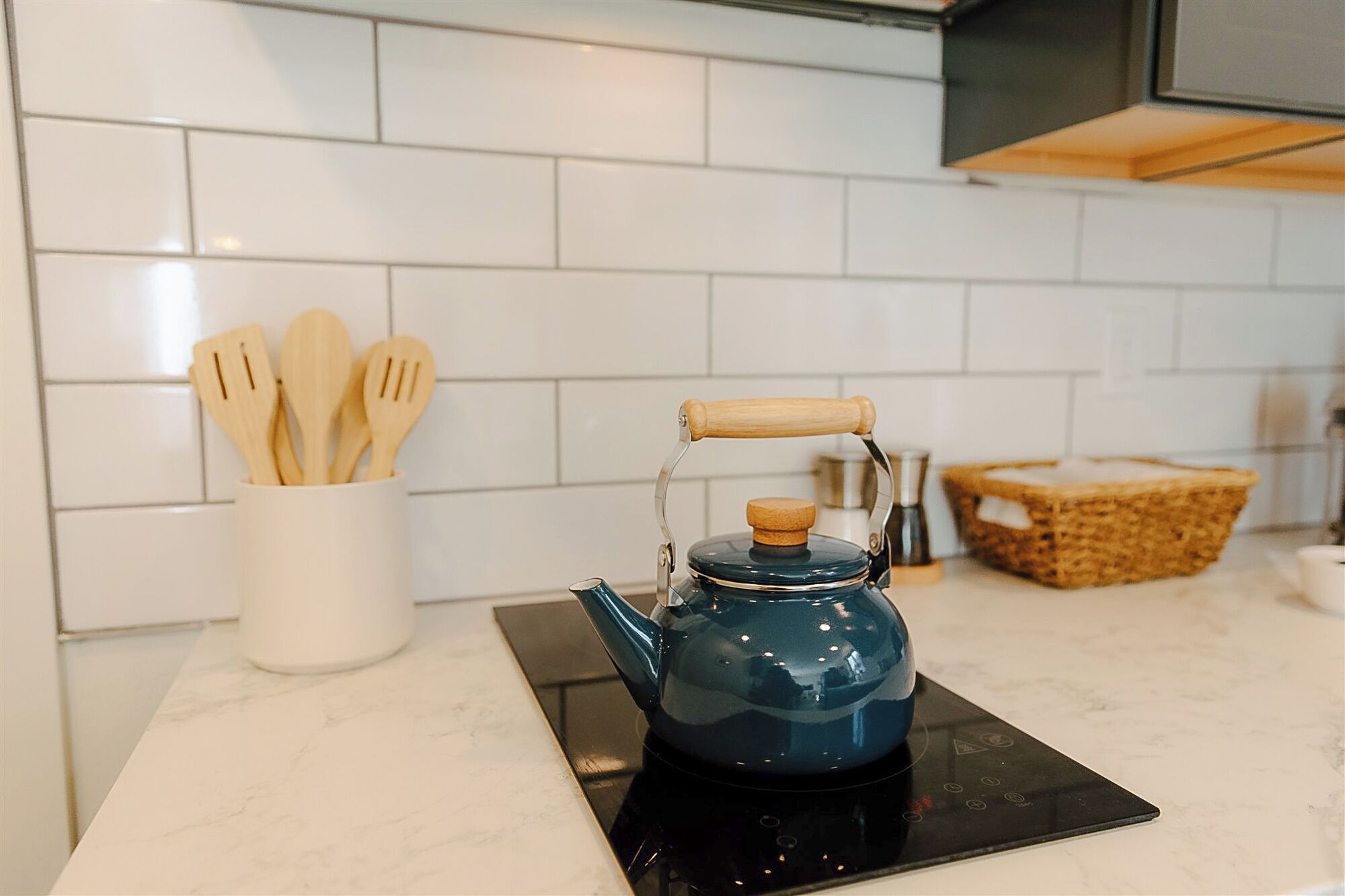 Uma cozinha com uma chaleira azul sobre um fogão por indução, utensílios de madeira em suporte branco e uma cesta de palha no balcão.