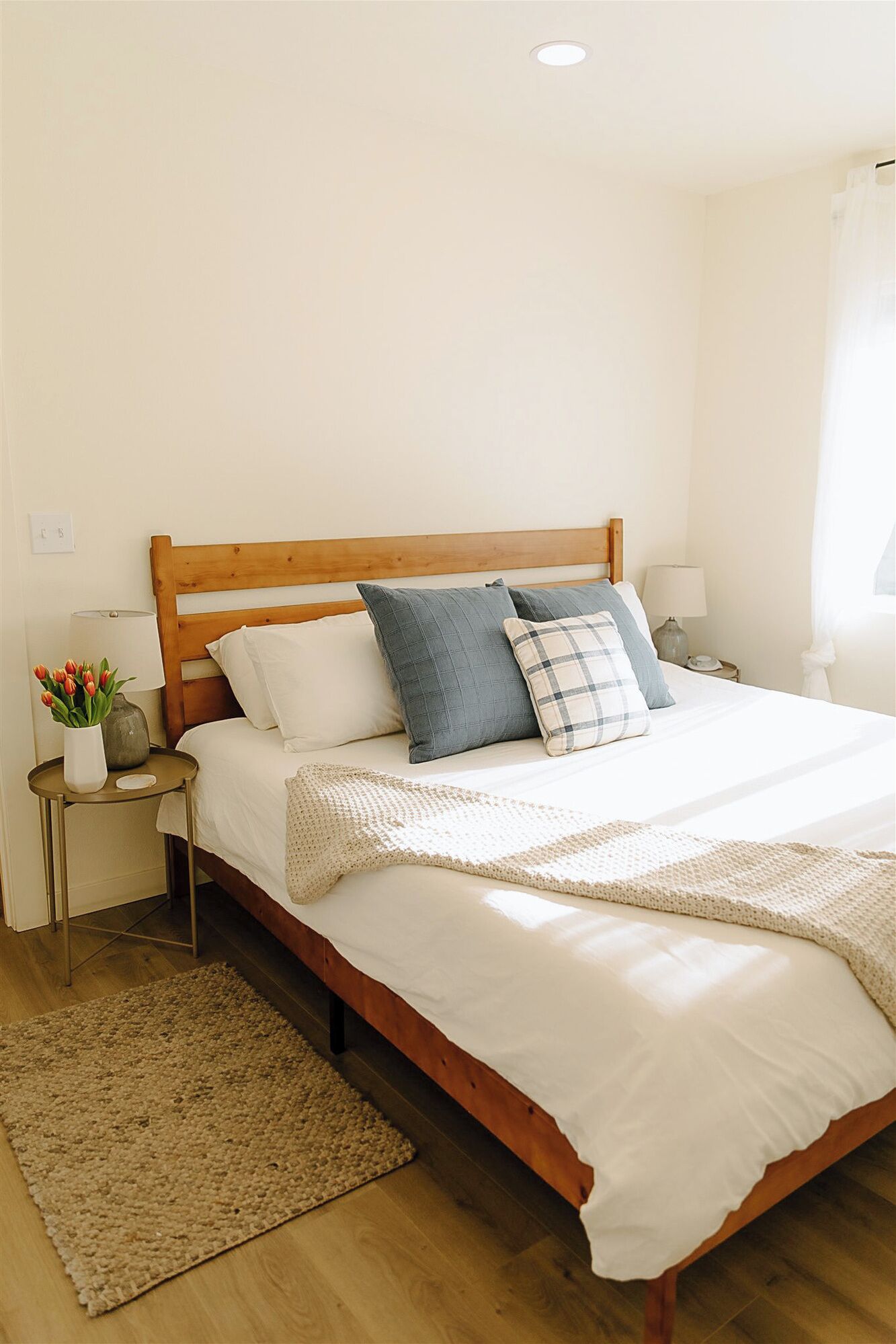 A neatly arranged bedroom with a wooden bed frame, white bedding, and a small bedside table with a lamp and a vase of flowers.
