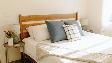 A neatly arranged bedroom with a wooden bed frame, white bedding, and a small bedside table with a lamp and a vase of flowers.