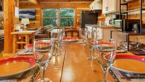A wooden dining table set with wine glasses and plates, a sofa, and a kitchen area with cabinets and appliances. - Pet-Friendly Townsend Mtn Cabin w/ Hot Tub & Daybed Patio (Townsend)