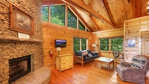 A log cabin interior with a stone fireplace, wooden walls, a sofa, and a television. - Pet-Friendly Townsend Mtn Cabin w/ Hot Tub & Daybed Patio (Townsend)