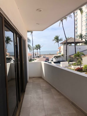 A balcony with a view of the beach, palm trees, and a building with a thatched roof. - The Best Beachfront Casita in Mazatlán for Rent! (Mazatlán)