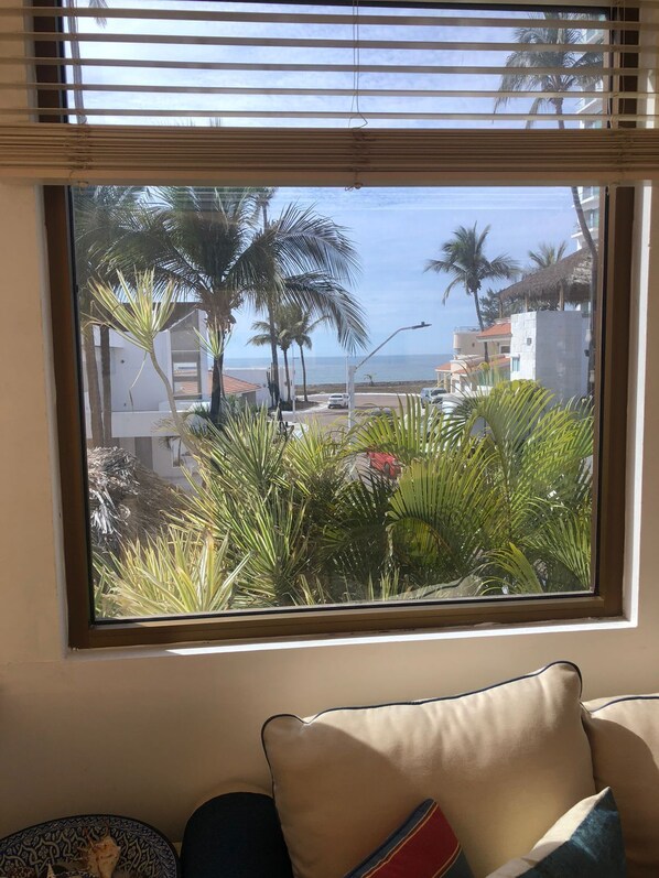 A window with a view of palm trees and buildings, a sofa with cushions, and a ceiling fan. - The Best Beachfront Casita in Mazatlán for Rent! (Mazatlán)