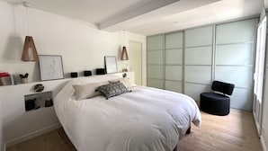 A modern bedroom with a large bed, a bedside table, a chair, a wardrobe, and a window.