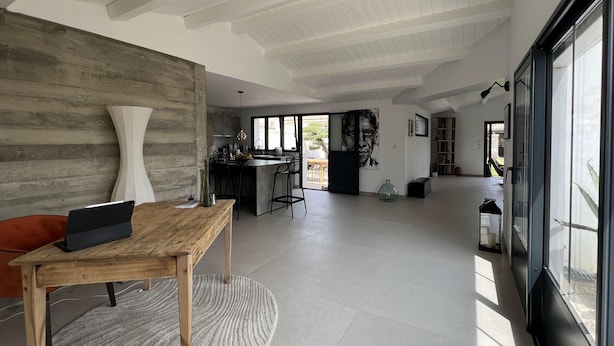A spacious living area with a wooden dining table, a large white vase, and a modern kitchen in the background.