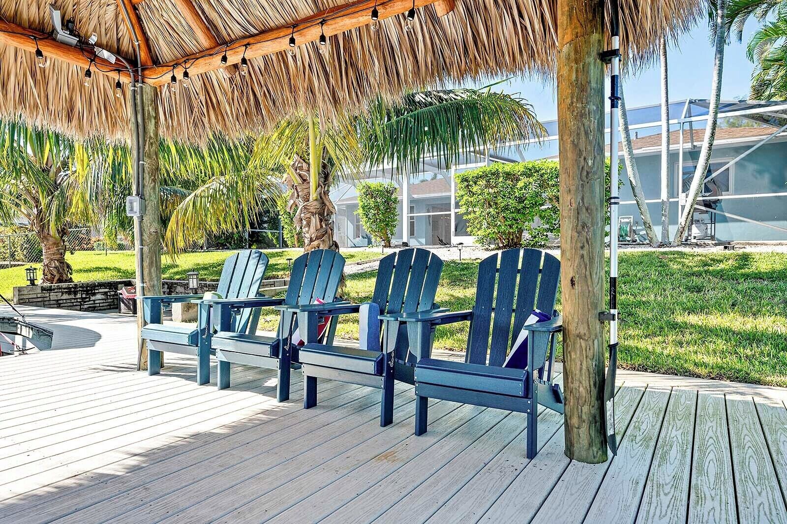 A wooden deck with three Adirondack chairs under a thatched roof, surrounded by greenery and a modern building in the background.
