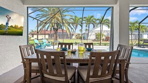Un coin repas avec une table et des chaises, une grande fenêtre donnant sur des palmiers et une maison, et un tableau représentant un golfeur accroché au mur.
