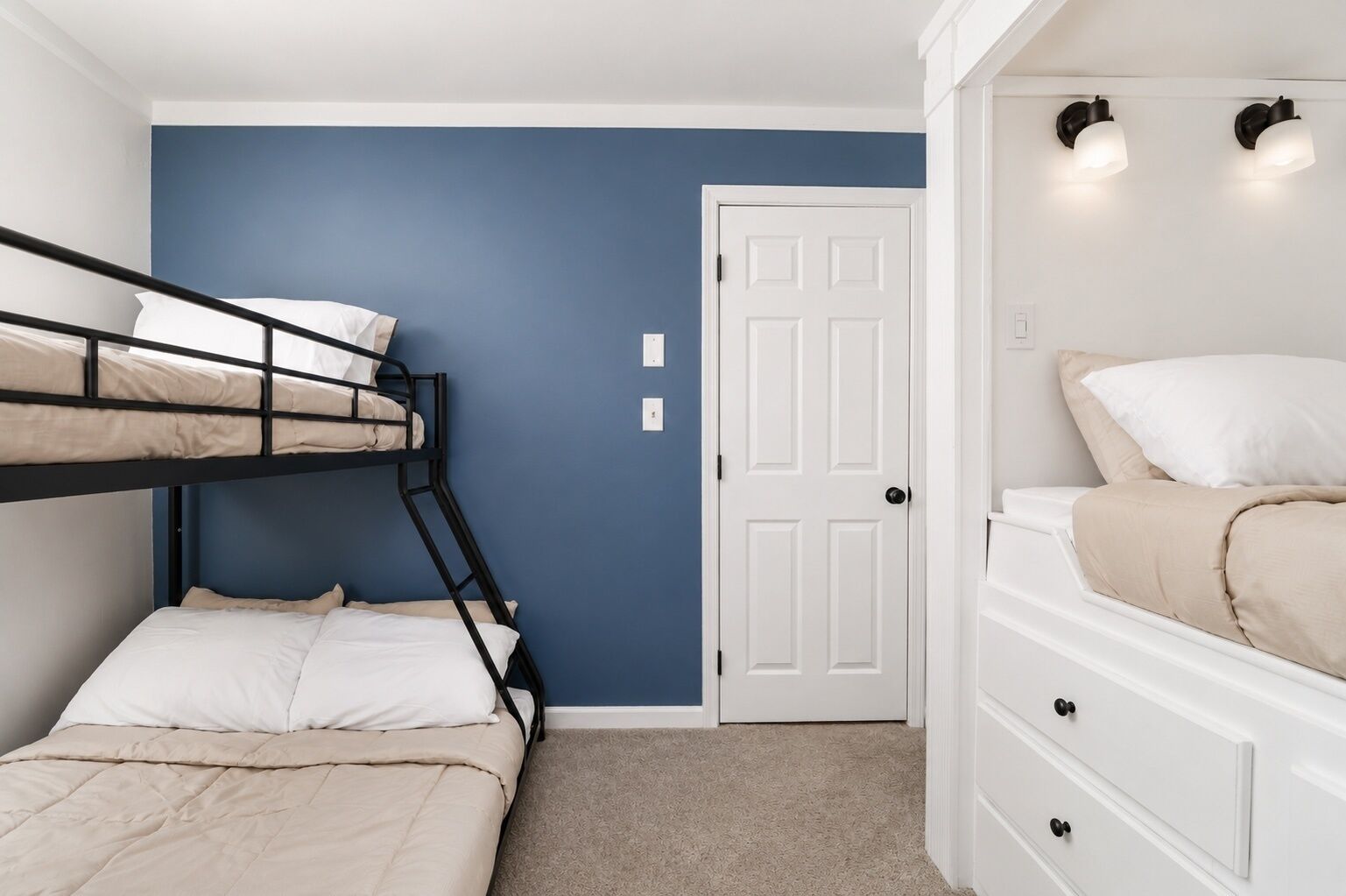 A room with bunk beds, a white dresser, a door, and wall-mounted lights.